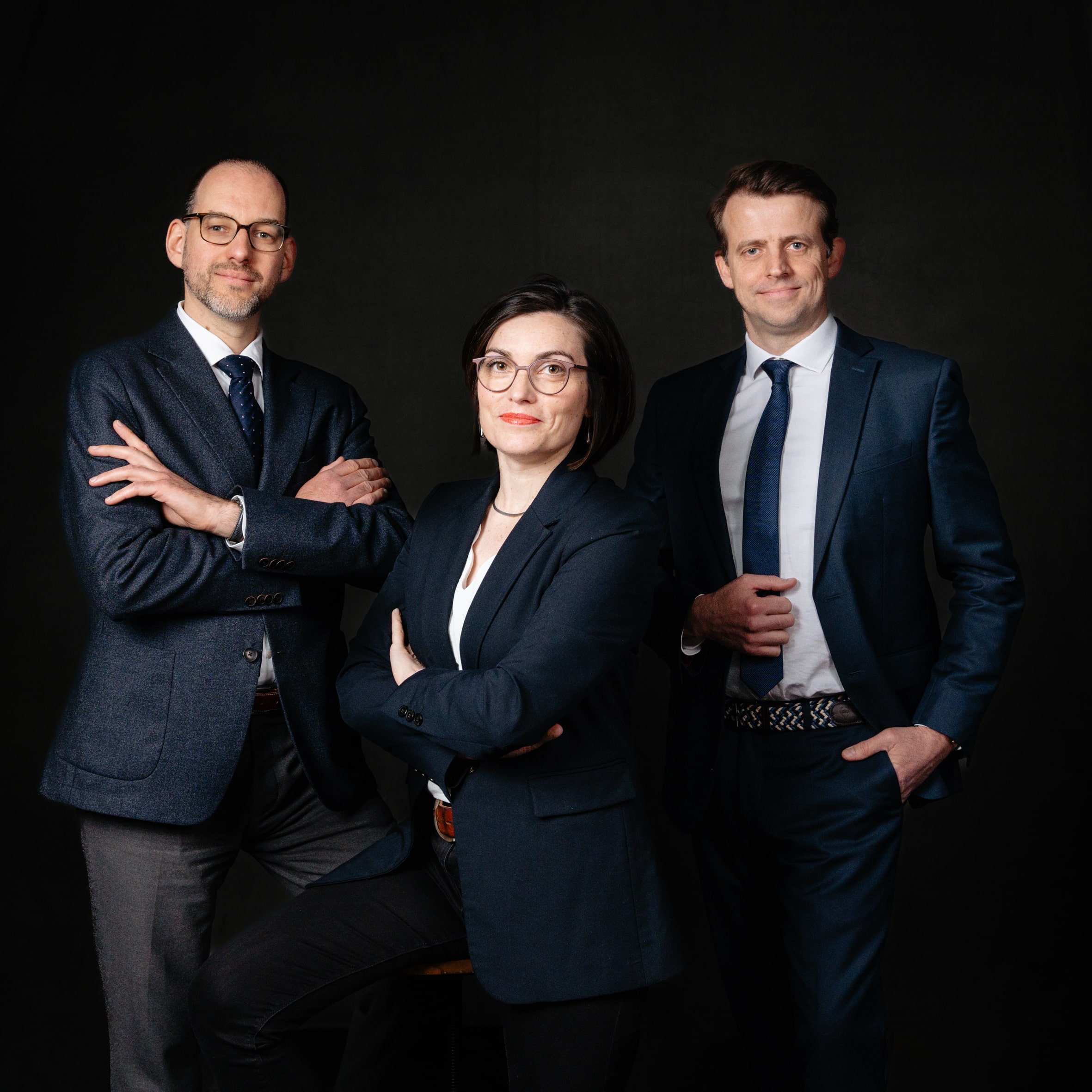 **Texte alternatif :**</p>
<p>Un portrait de groupe professionnel montrant trois avocats posant ensemble, capturé par Frédérique Jouvin. Les deux hommes et la femme, tous vêtus de costumes élégants, affichent une posture confiante et professionnelle. L'homme à gauche porte un costume gris foncé avec une cravate assortie, la femme au centre est habillée d'un blazer noir et de lunettes, et l'homme à droite porte un costume bleu marine avec une cravate bleue. Ils se tiennent devant un fond sombre, ce qui met en valeur leur apparence formelle et leur présence dynamique. Ce portrait reflète l'image d'une équipe cohésive et compétente, prête à représenter leurs clients avec professionnalisme et assurance.